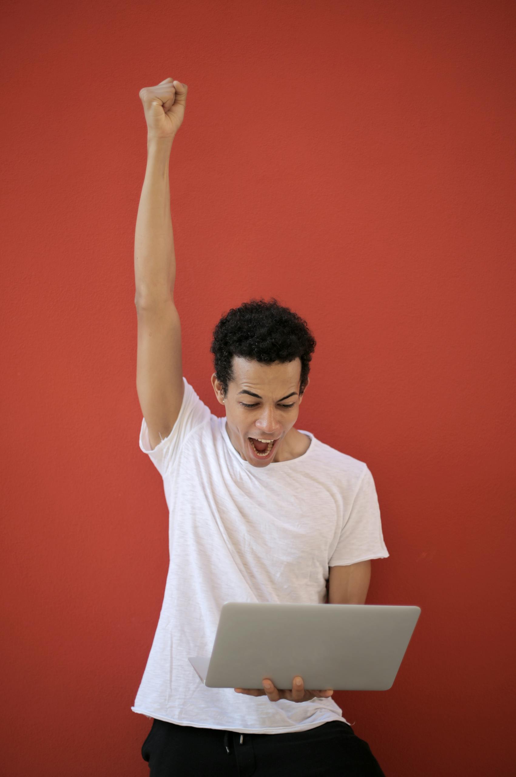 A young man in a white shirt celebrates success holding a laptop against a red background.